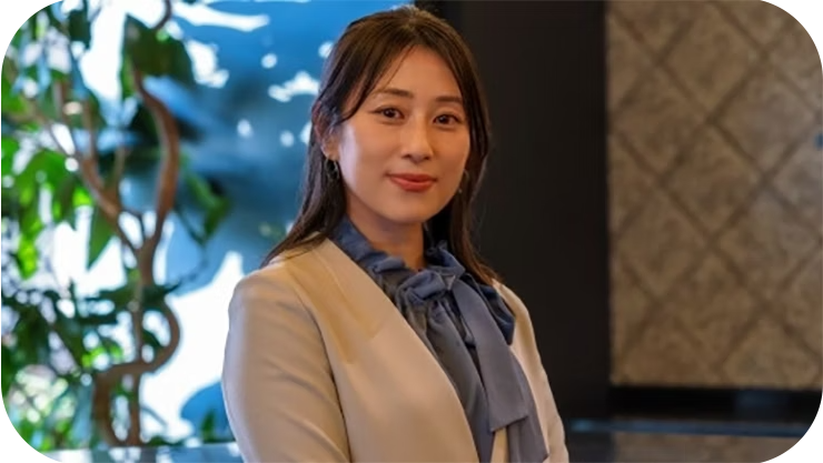 チサはクリーム色のブレザーを着て室内に立っており、背景には葉の茂った植物と柔らかな光に照らされた壁が見える。