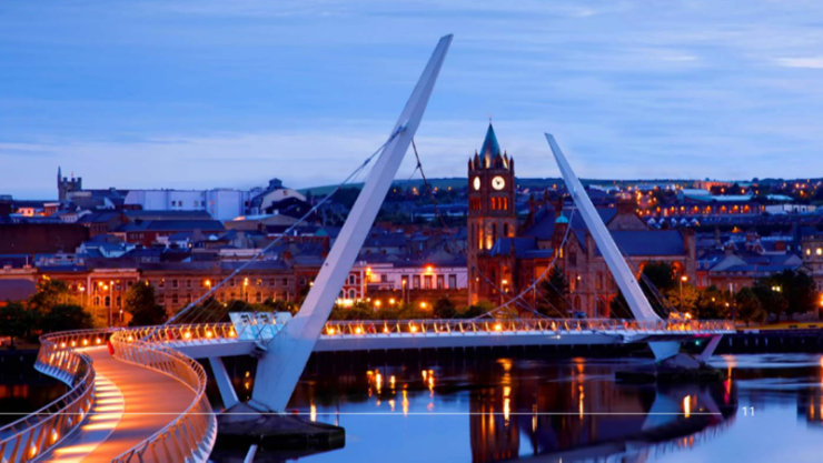 The Peace Bridge over the River Foyle in Derry-Londonderry at dusk, with the illuminated Guildhall clock tower and city skyline reflecting on the water. The number 11 appears in the bottom right corner.