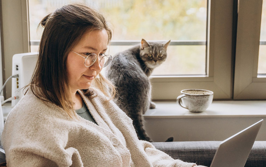 Young woman working from home uses a laptop while sitting on the sofa with her cat.