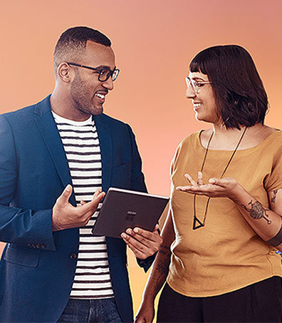 Eine Frau und ein Mann diskutieren vor einem farbigen Hintergrund und lächeln. Der Mann hält ein Tablet