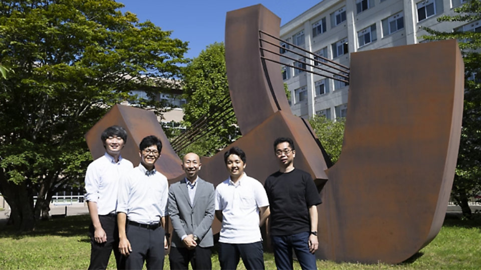 笑顔の男性5人が屋外で、大きな抽象的な金属彫刻と建物を背景にポーズをとっている。