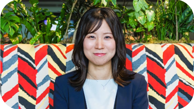 友美は、明るい色のトップスの上に紺色のブレザーを羽織り、カラフルなジグザグ模様のベンチの前に座っている。彼女の後ろには緑の植物が植えられている。