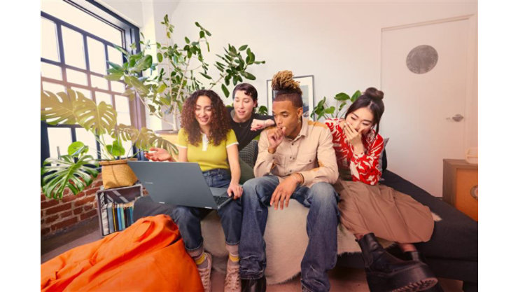 明るい植物のある部屋のソファに座った4人の若者がノートパソコンを見ています。一人の女性が身振り手振りで話し、もう一人は口を覆い、男性が見ています。