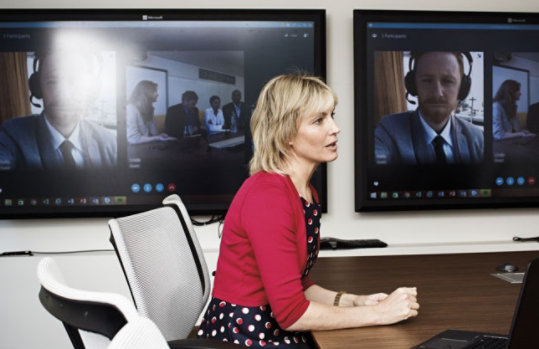 Eine Frau sitzt in einem Konferenzraum und hält eine Videobesprechung in Microsoft Teams. An der Wand hängen zwei große Bildschirme.