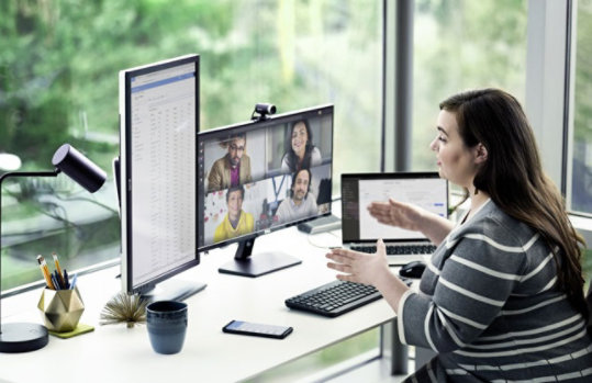 Eine Frau sitzt an einem Schreibtisch mit drei Bildschirmen und führt eine Videobesprechung in Microsoft Teams mit vier Kollegen.