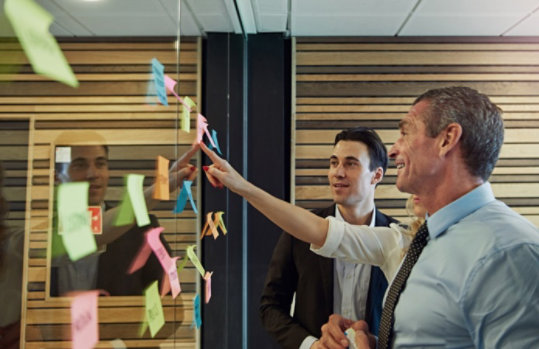 Kreativmeeting, in dem eine Frau zwei Männern Post-its an der Wand zeigt