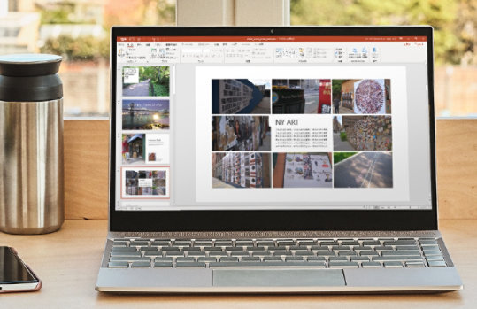 木製の机の上に置かれたノートパソコンが、「NY ART」と題された画像付きのPowerPointスライドを表示しており、隣にはボトル、スマートフォン、メガネが置かれています。