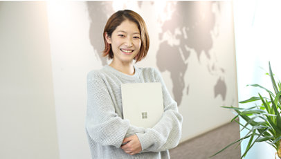 灰色のセーターを着た笑顔の女性がオフィスでマイクロソフトのノートパソコンを持っています。