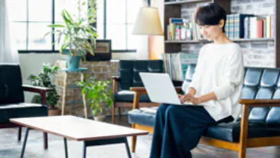 ソファでノートパソコンを使う人、明るい部屋に植物と本棚。