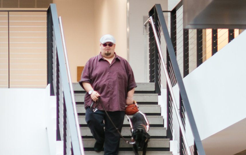 Ein Mann, der eine Sehbehinderung hat, wird von einem Hund begleitet, während er die Stufen einer Treppe hinuntergeht