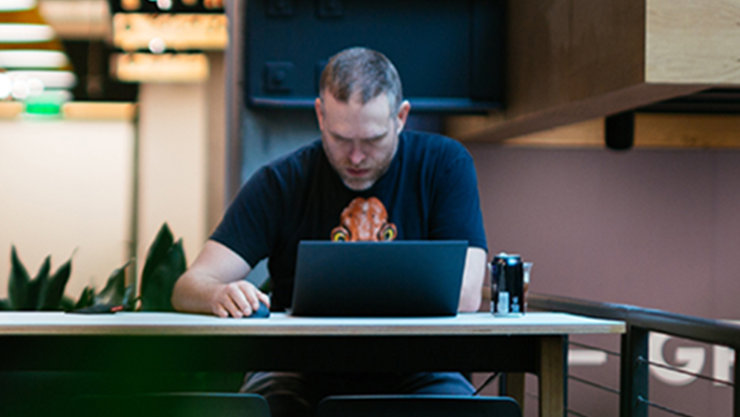 Ein Mann sitzt frontal zur Kamera und sieht konzentriert auf seinen Laptop, während er eine Maus bedient