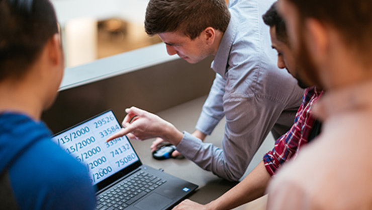 Ein Mann deutet auf einen Laptop mit vergrößerter Tabellenansicht, während er in einer Menschengruppe sitzt