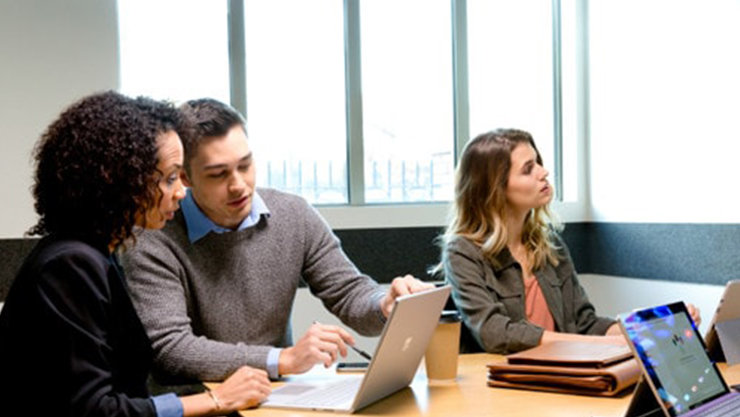 Zwei Personen sitzen an einem Tisch und blicken in einen Laptop, während eine dritte Person neben ihnen sitzt.