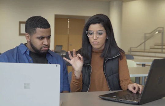 Zwei Personen sitzen an einem Schreibtisch, beide haben jeweils einen Laptop vor sich aufgeklappt. Eine Person führt ein Zeichen der Gebärdensprache aus, während die andere Person auf den anderen Laptop sieht.