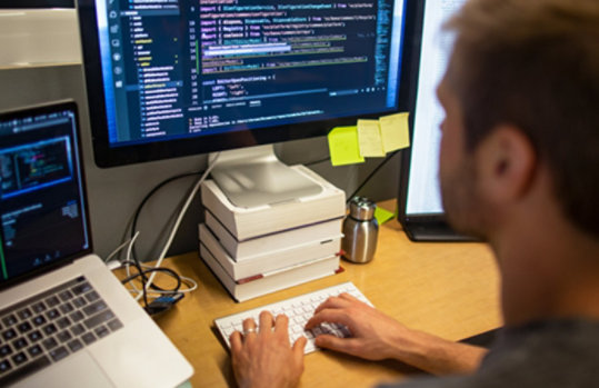 男性開発者は、MacBook Pro のマルチモニター設定で Visual Studio を使用して、オフィスのワークスペースでコーディングしています