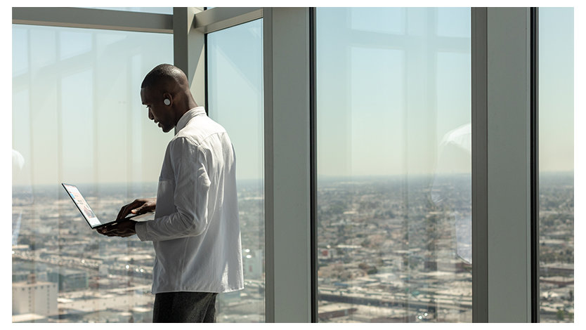 高層ビルの窓際に立って開いたノート PC を操作するビジネス パーソン
