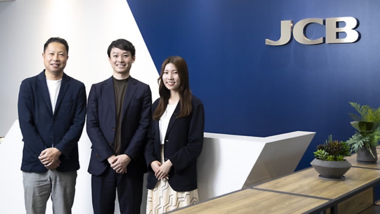 JCBのロゴがある青い壁の前で、3人のビジネスパーソン（男性2人、女性1人）が立っている。