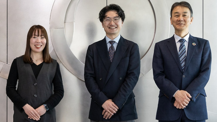 白い壁の前に立つ3人の人物（女性1人、男性2人）。皆笑顔でカメラを見ている。