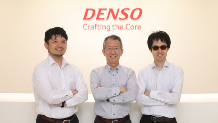 DENSOのロゴを背景に、腕を組んで笑顔で立つ3人の男性。