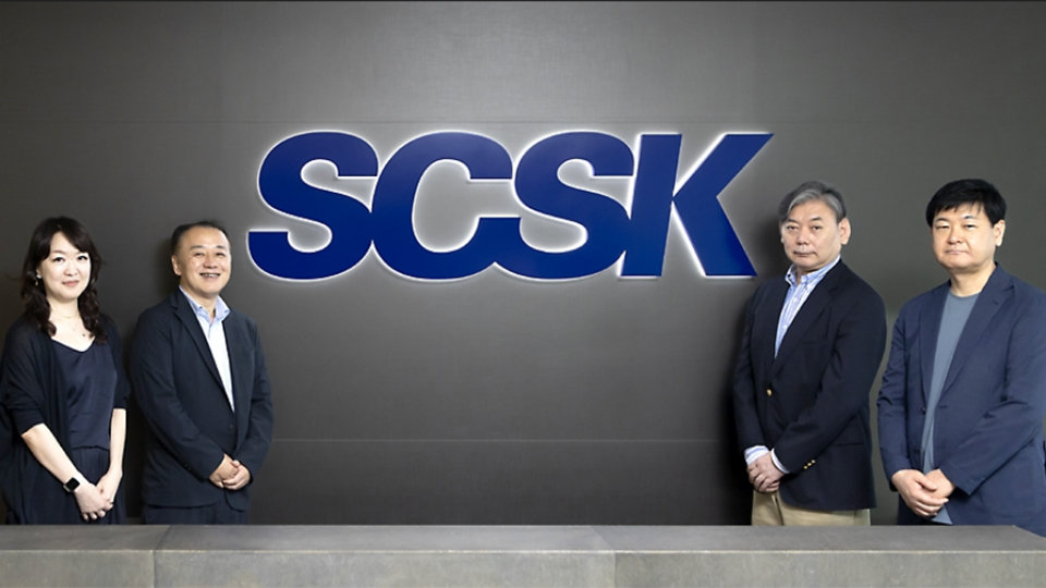 「SCSK」のロゴを背景に、4人のビジネスパーソンが立っている。