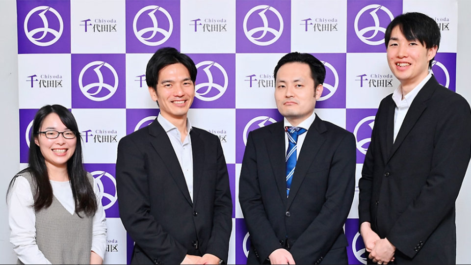 千代田区のロゴが入った紫と白の背景の前で、笑顔の4人の人物（女性1人、男性3人）が立っている。