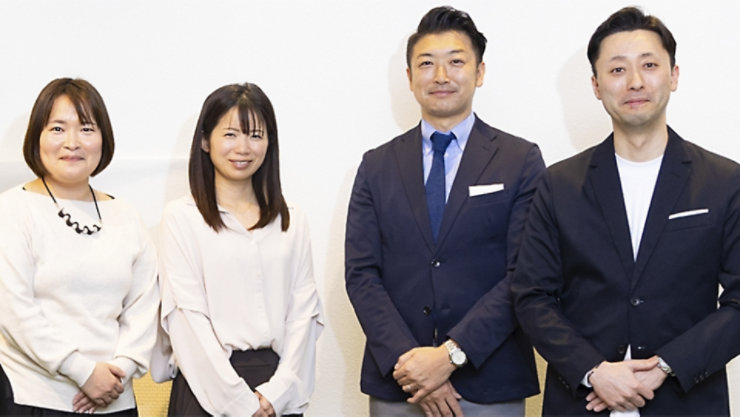 白い背景の前で、2人の女性と2人の男性が立っており、皆カメラに向かって微笑んでいる。