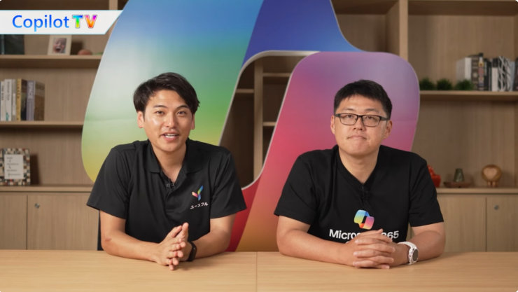 THINKYのロゴとMicrosoft 365のロゴが入ったTシャツを着た2人の男性が、カラフルな背景の前で机に座っている。