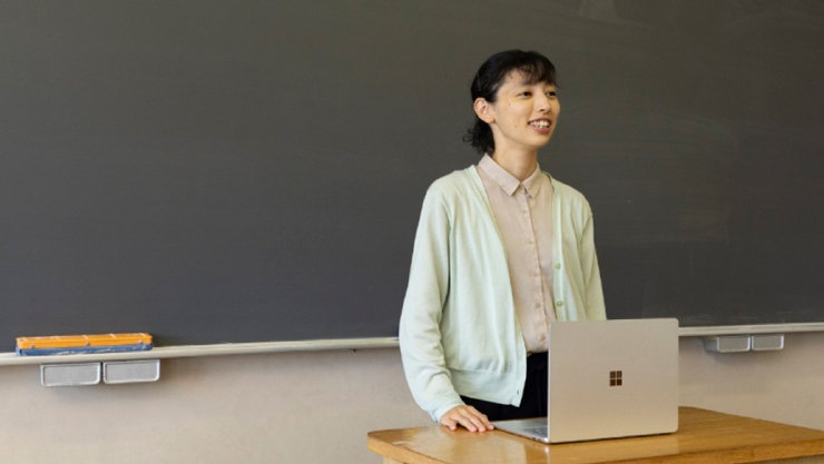笑顔で黒板の前に立つ女性。木製の机にはMicrosoft SurfaceノートPCが開いて置かれている。