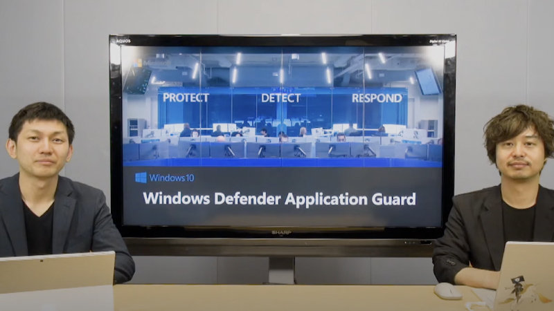 男性2人がテーブルに座り、背後には「Windows Defender Application Guard」と書かれたモニターがある。