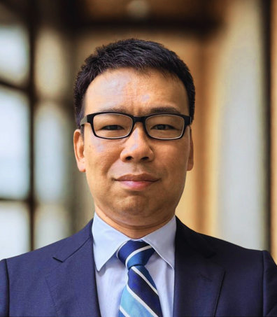 中村 弘毅氏が、背景のぼけた室内でスーツとストライプのネクタイを着て立っている。