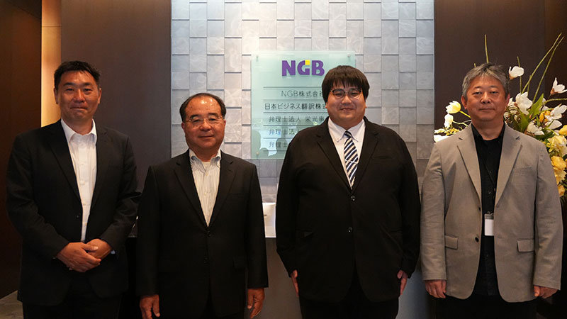 「NGB」の看板の前で、屋内に並んで立つビジネス attire の4人。