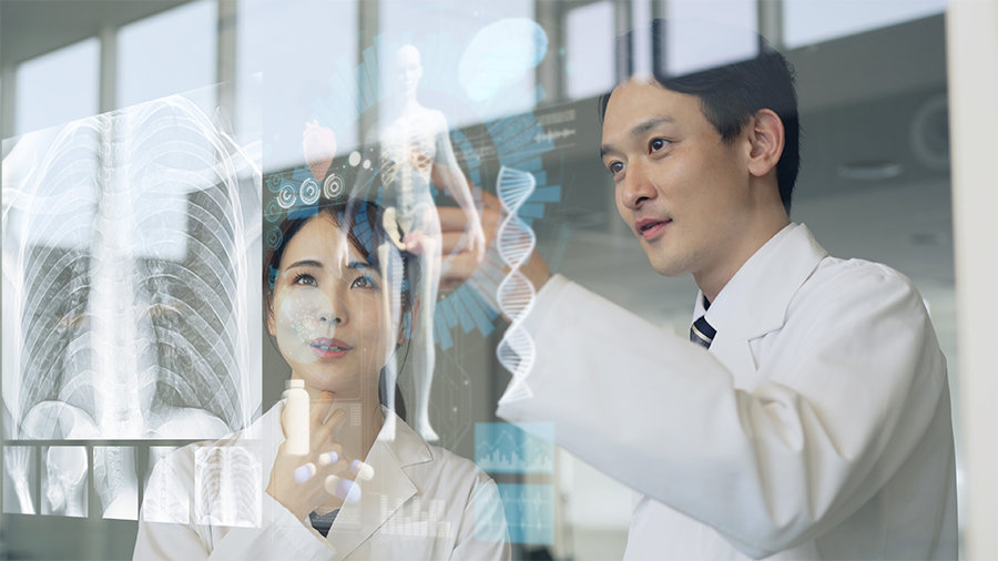 透明なボードに映し出された人体の画像を見ながら話す白衣を着た 2 人の人物