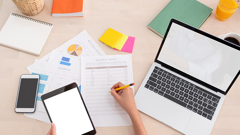 ある人物が財務データを確認し、メモを取っています。作業スペースには、ノートパソコン、タブレット、電話、印刷されたグラフや表が含まれます。