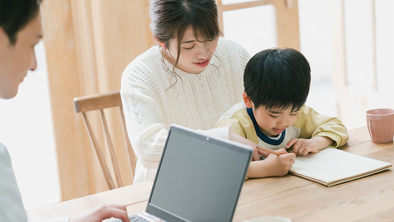 ダイニングテーブルでノート PC で仕事をする父親とその前に座る母親と子ども