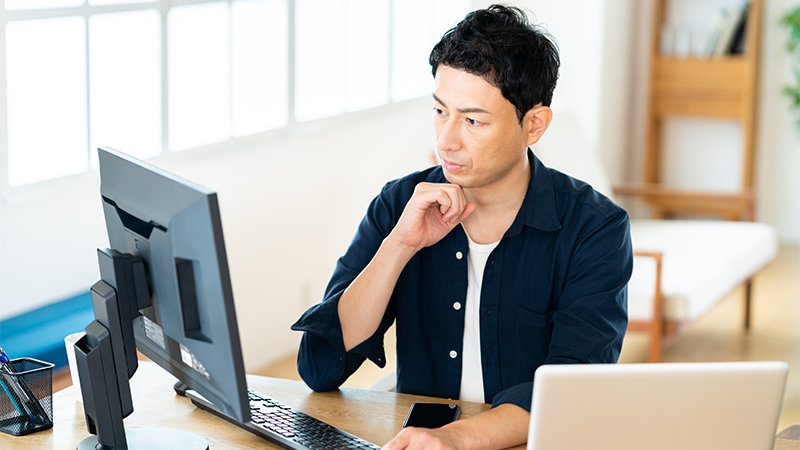 男性が机に座り、考え込むように顎に手を当てて、コンピューターのモニターを見ています。机の上にはノートパソコンとスマートフォンも置いてあります。背景には明るい窓と棚が見えます。