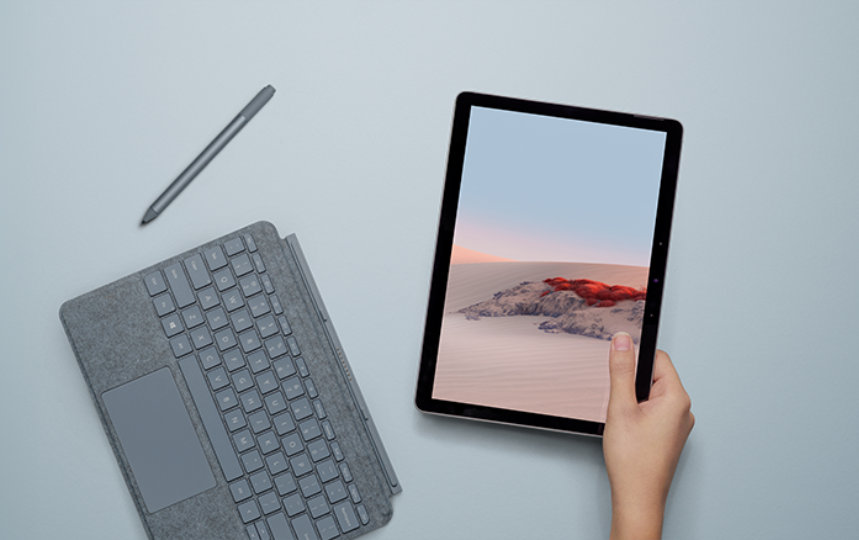 Surface ペンと Surface Go タイプカバーが近くに置かれた机の上で、Surface Go 2 を持つ人