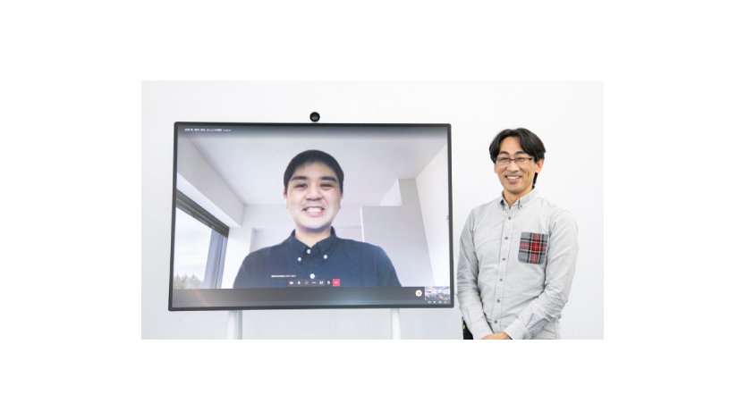 大きなモニターの横に立って別の男性を映す笑顔の男性