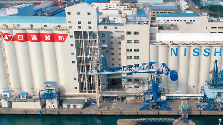 港湾に立地する日清製粉工場の俯瞰。白いサイロに赤い「日清製粉」ロゴと青い「NISSHIN」の文字、岸壁には青色のクレーンが並ぶ。