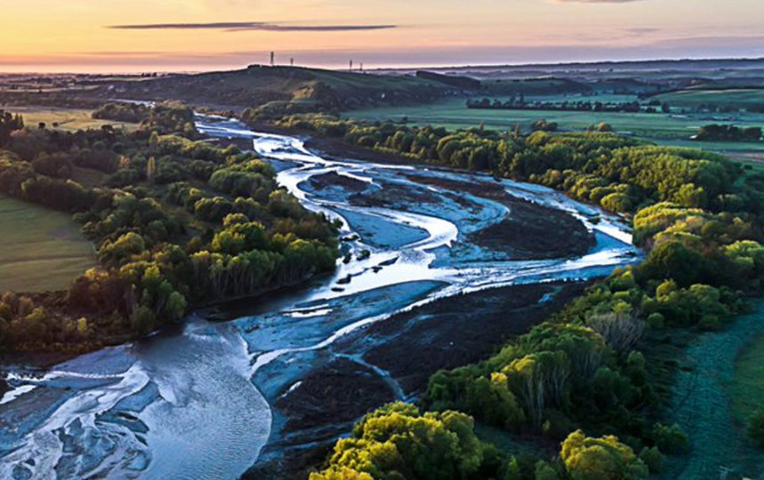 An aerial view of a wide, braided river flowing through a lush green valley at sunset. The sun is setting on the left, casting a bright golden glow over the landscape. Green fields and dense trees line the winding river, with rolling hills visible in the distance.