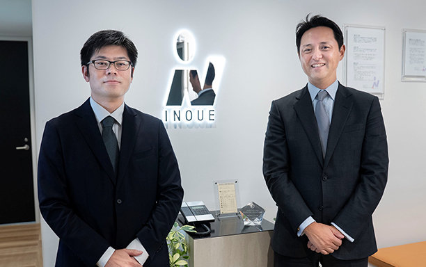 受付カウンター前でスーツ姿のビジネスパーソン2名が立ち、背後にINQUEのロゴがあるオフィス風景。