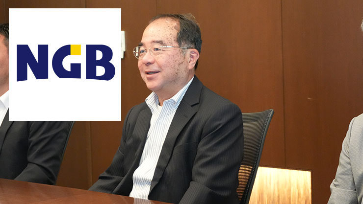 会議テーブルに座り、右向きに話すダークカラーのブレザーと淡色のシャツを着た人物。左側のパネルにNGBロゴが表示され、背景は木製の壁