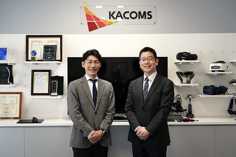 「KACOMS」のロゴが掲げられたオフィスで、スーツ姿で笑顔を見せる2人の男性