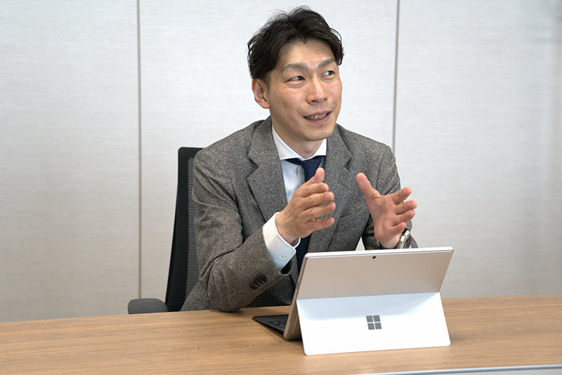 タブレットPCを前に置き、ジェスチャーを交えながら笑顔で話すグレーのジャケット姿の男性。