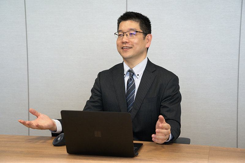 ノートPCを前に置き、両手を広げるジェスチャーをしながらにこやかに話すスーツと眼鏡姿の男性。