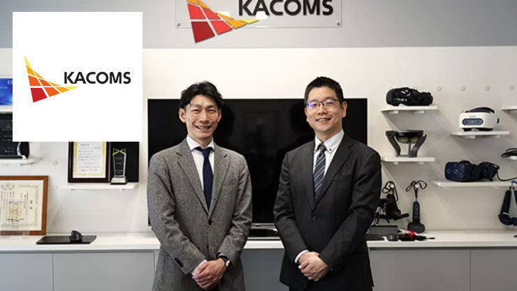 「KACOMS」のロゴマークを背に、笑顔で並んで立つスーツ姿の男性2人。背景の棚にはVRヘッドセットや表彰盾がディスプレイされています。