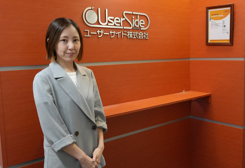 株式会社ユーザーサイド　営業推進課　課長　廣川 唯 氏オレンジ色の壁の前に立つ"UserSide SAPPORO | ユーザーサイド株式会社" logos.