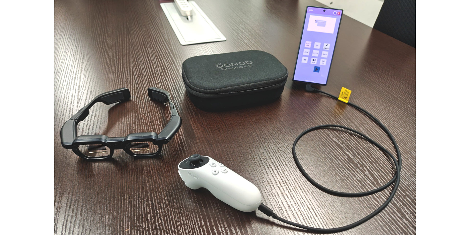 QONOQ Devicesのケース、黒いスマートグラス、白いコントローラー、接続されたスマートフォンが並ぶテーブル。