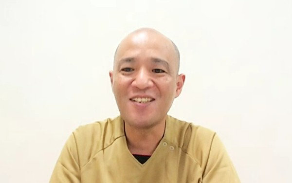 ベージュのスクラブを着て笑顔で正面を向くスキンヘッドの男性。