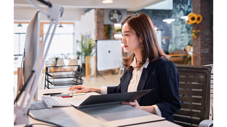 ビジネスウェアに身を包んだ女性が、コンピューターのキーボードや書類、オフィス用品が置かれたモダンなデスクで仕事に取り組んでいます。自然光や観葉植物、開放的なワークスペースのデザインが取り入れられたこの環境は、プロフェッショナルかつ整然とした職場環境であることを示唆しています。