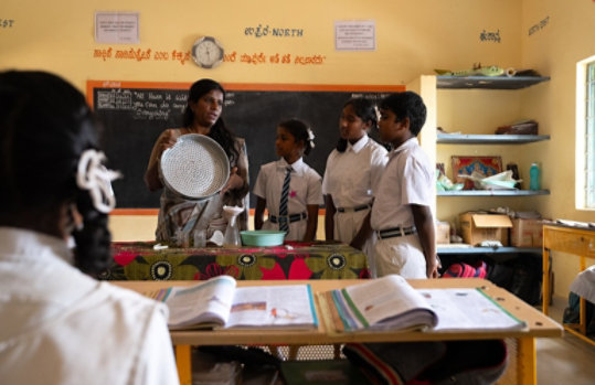 An Indian school classroom where a female teacher in traditional attire holds a metal sieve, explaining to attentive uniformed students near a decorated table with educational materials, with an open textbook in the foreground.